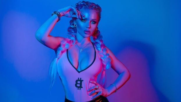 Fashion portrait of stunning sexy young blonde woman with trendy braids and sparkling bra under the net top. She is holding peace sign by the eye, making pouting lips in bright blue and red light.