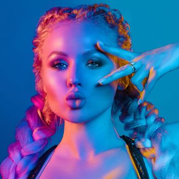 Fashion portrait of stunning sexy young blonde woman with trendy braids and sparkling bra under the net top. She is holding peace sign by the eye, making pouting lips in bright blue and red light