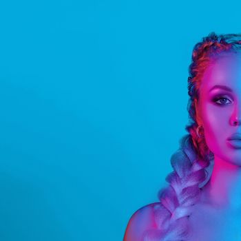 Fashion portrait of stunning sexy young blonde woman with trendy braids and sparkling bra under the net top. She is holding peace sign by the eye, making pouting lips in bright blue and red light. Studio fashion portrait.