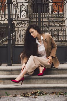 Close up portrait of young beautiful woman with long brunette curly hair posing against building background. woman wearing a white blouse and beige pants and a jacket