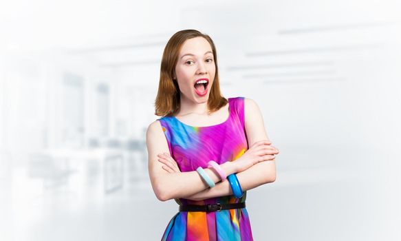 Beautiful caucasian woman standing with folded arms. Elegant fashionable lady in colorful dress and bracelets. Studio shot of good looking redhead girl with open mouth on blurred office background