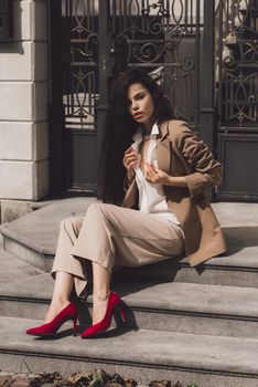 Close up portrait of young beautiful woman with long brunette curly hair posing against building background. woman wearing a white blouse and beige pants and a jacket