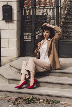 Close up portrait of young beautiful woman with long brunette curly hair posing against building background. woman wearing a white blouse and beige pants and a jacket