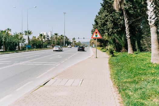 Traffic light sign standing on the pavement near the highway in the southern city with palms on the sideline. Stop light sign near the busy road