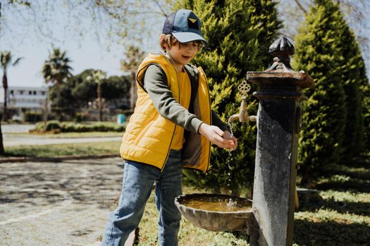 Kid schoolboy in casual clothing washing his hands in the street old fashioned drinking water fountain. Boy in blue jeans and yellow vest playing with water from drinking sprinkler in city park.