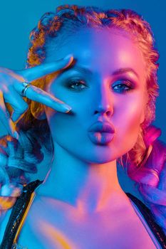 Fashion portrait of stunning sexy young blonde woman with trendy braids and sparkling bra under the net top. She is holding peace sign by the eye, making pouting lips in bright blue and red light