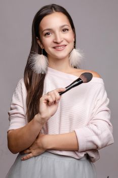 Beautiful cute girl in sweater with one naked shoulder posing at camera. Young make up artist holding one hand on belly and in other big brush. Happy brunette lady with pink feather earrings smiling.