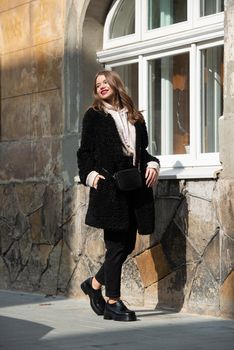 Fashion photo of young woman in black fur coat, jeanse and shoes at city street. beige jacket with a hood