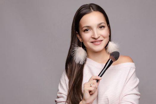 Beautiful cute girl in sweater with one naked shoulder posing at camera. Young make up artist holding one hand on belly and in other big brush. Happy brunette lady with pink feather earrings smiling.