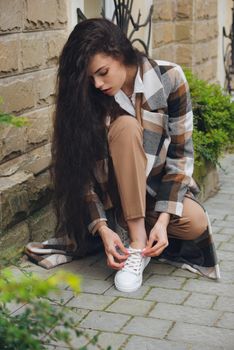 Closeup portrait of young beautiful fashionable woman wearing checkered long coat, beige pants and white blouse . Women ties laces on sneakers. Female fashion.