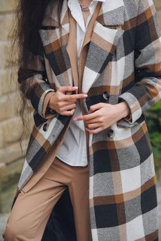 Closeup portrait of young beautiful fashionable woman wearing checkered long coat, beige pants and white blouse . Lady posing on city street.. Female fashion.