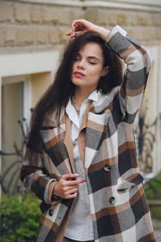 Closeup portrait of young beautiful fashionable woman wearing checkered long coat, beige pants and white blouse . Lady posing on city street.. Female fashion.