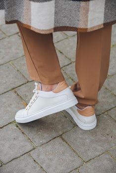 Closeup photo of beautiful fashionable woman wearing checkered long coat, beige pants, white blouse and sneakers. Lady posing on city street.