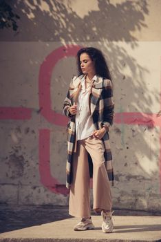 Closeup portrait of young beautiful fashionable woman wearing checkered long coat, beige pants and white blouse . Lady posing on city building background. Female fashion.