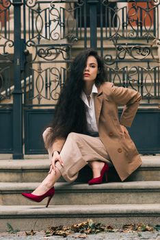 Close up portrait of young beautiful woman with long brunette curly hair posing against building background. woman wearing a white blouse and beige pants and a jacket