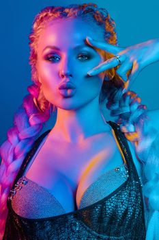 Fashion portrait of stunning sexy young blonde woman with trendy braids and sparkling bra under the net top. She is holding peace sign by the eye, making pouting lips in bright blue and red light