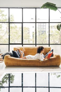 Young mixed race woman reading a book at home living room. Copy space. Vertical image. Lifestyle concept.