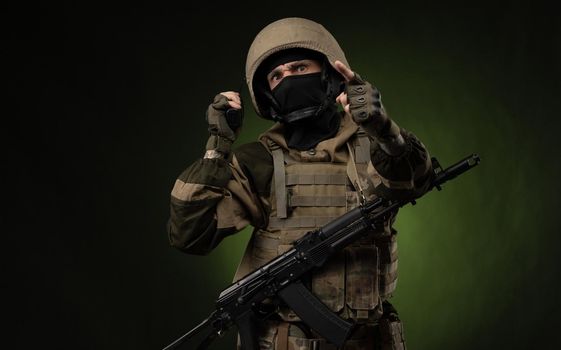 Russian soldier in military clothes with a Kalashnikov assault rifle on a dark background takes orders on a walkie-talkie