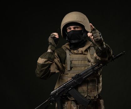 Russian soldier in military clothes with a Kalashnikov assault rifle on a dark background takes orders on a walkie-talkie