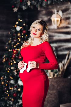 View of beautiful blonde pregancy mom lying, posing at studio, looking, smiling at camera. Mother expacting for child, posing at decorated studio for new year and Christmas holiday. Woman wearing in white for, with make up for x mas paty.