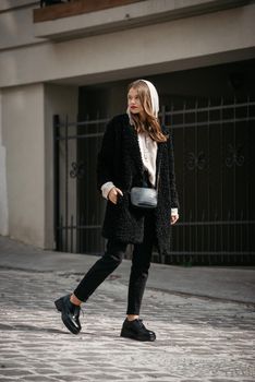 Fashion photo of young woman in black fur coat, jeanse and shoes at city street. beige jacket with a hood