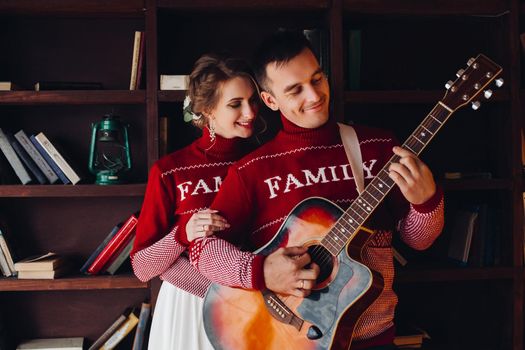 Beautiful smiling couple in red sweaters listening music, girlfriend leaning on handsome boyfriend, when he playing at gitar. Celebrating new year and xmas. Romantic winter holidays.