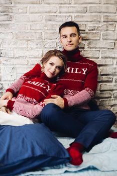 Portrait of young positive couple wearing in red sweaters with discription family. Happy boyfriend and girlfriend embracing and leaning on wall at studio. Wife and husband celebrating xmas.