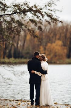 Back view of beautiful young bride in sweater and dress and groom in black coat and suite standing at river or lake pear, embracing and kissing. Beloved couple walking, celebrating wedding at autumn.