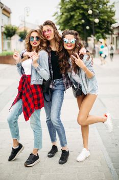 Full length of stylish trendy girlfriends in mirrored sunglasses and summer clothes posing embracing in the street.
