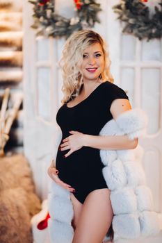 Portrait of pregnancy blonde woman looking, smiling at camera expecting little child, embracing her stomach. Mom posing at studio decorated for new year and Christmas, wearing in fur. Holliday.