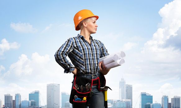 Attractive female engineer in hardhat standing with technical blueprints. Portrait of young architect in checkered blue shirt on background of cityscape. Architecture and construction company.