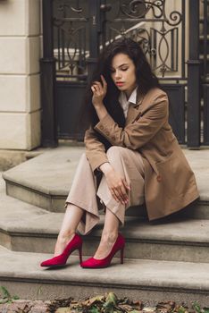 Close up portrait of young beautiful woman with long brunette curly hair posing against building background. woman wearing a white blouse and beige pants and a jacket