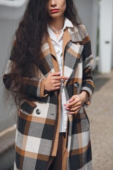 Closeup portrait of young beautiful fashionable woman wearing checkered long coat, beige pants and white blouse . Lady posing on city street.. Female fashion.