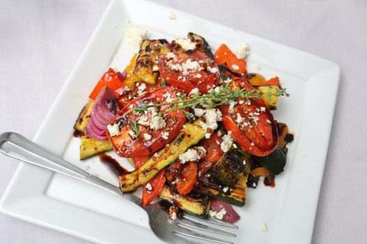 Grilled Vegetables with Tomatoes Peppers Zucchinis Red Onion and Fresh Thyme Sprig Drizzled with Balsamic Vinegar and Crumbled Feta Cheese on Square White Plate on White Tablecloth. High quality photo