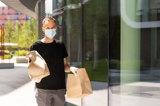 Delivery guy with protective mask and gloves holding bag with groceries in front of a building