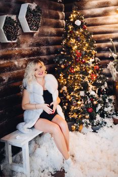 Pretty and young pregnancy mom sitting on bench in stylish decorated studio with new year tree in corner and snow around. Blonde mother holding hands on stomach, showing heart by fingers, smiling at camera. Expacting baby.