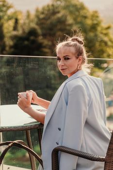 A middle-aged woman sits in a street cafe overlooking the mountains at sunset. She is dressed in a blue jacket and drinks coffee admiring the nature. Travel and vacation concept