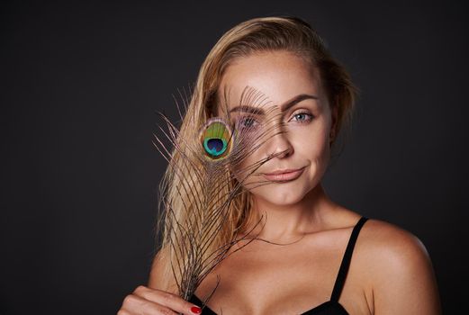 Close-up studio portrait of a beautiful blonde Caucasian woman holding a peacock feather isolated over black background with copy ad space