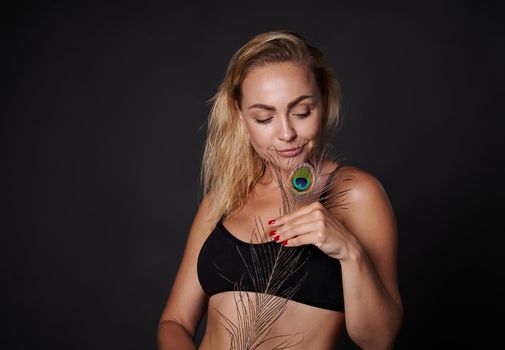 Attractive middle aged European woman in black underwear holding a peacock feather, posing against black background with copy ad space