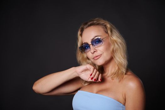 Confident portrait of a charming middle aged European woman with wavy blond hair wearing a blue top and blue sunglasses and looking at camera isolated over black background with copy ad space.