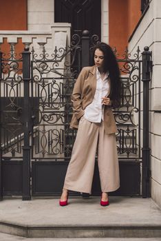 Close up portrait of young beautiful woman with long brunette curly hair posing against building background. woman wearing a white blouse and beige pants and a jacket