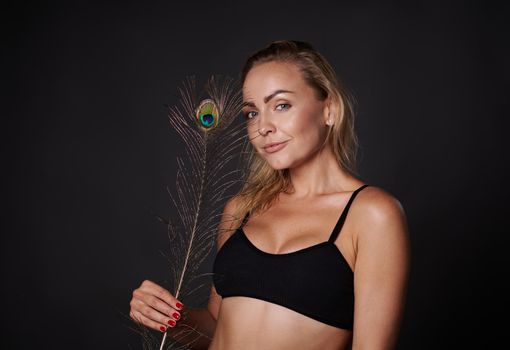 Attractive middle aged Caucasian woman in black underwear holding a peacock feather, posing against black background with copy ad space