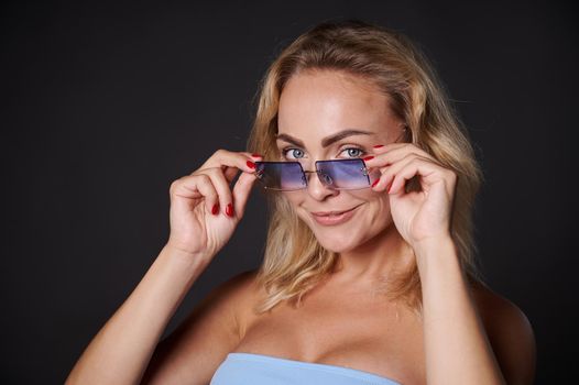 Studio portrait beautiful blonde Caucasian woman confidently looking at camera through blue sunglasses, isolated over black background with copy space for ads