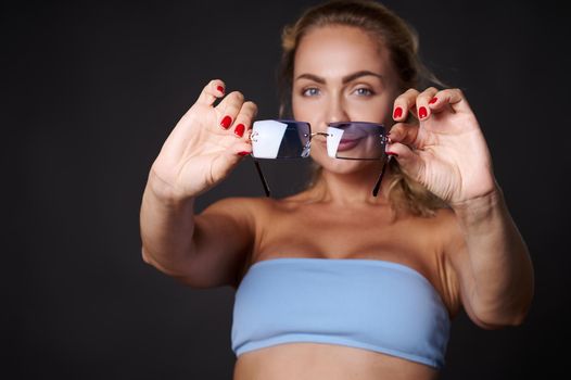 Attractive stunning beautiful blonde European woman confidently looking at camera through blue sunglasses, isolated over black background with copy space for ads