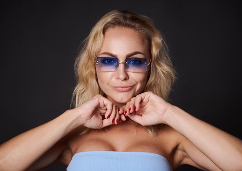 Studio portrait beautiful blonde Caucasian woman in blue sunglasses confidently looking at camera isolated over black background with copy space for ads