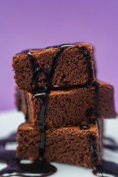 Very close shot of a Pile of Chocolate Brownies with chocolate strands falling on a white plate with purple background.