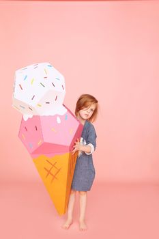 Portrait of a pretty little girl celebrating birthday and holding big ice cream toy in hands on over pink background.