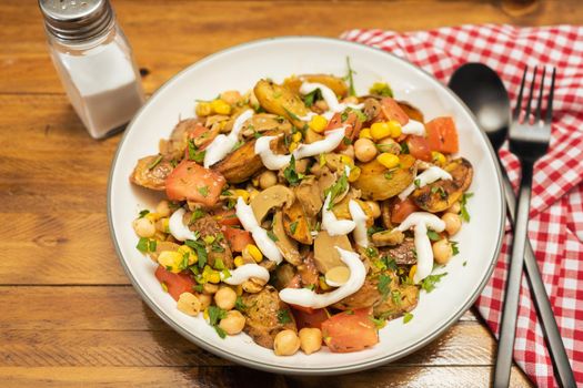 A delicious potato, chickpea, tomato and mushroom salad with parsley and aioli in a bowl with fork and spoon on a wooden table. Healthy, homemade, vegan food. High view.