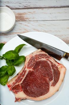 A bone-in steak or t-bone steak or porterhouse raw on a white plate on a wooden table. Top view. Vertical orientation. Close-up.