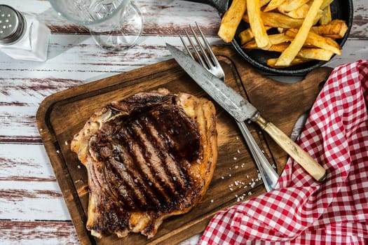 Incredible juicy t-bone cooked on the grill or barbecue on a wooden board accompanied by a french fries and with cutlery on the side. Top view.